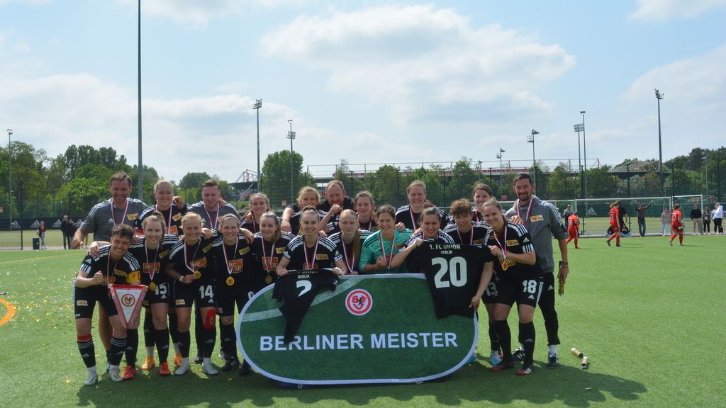 Mannschaft aus Frauenfußballspielern posiert auf dem Platz mit Medaillen und einem Banner „Berliner Meister“.