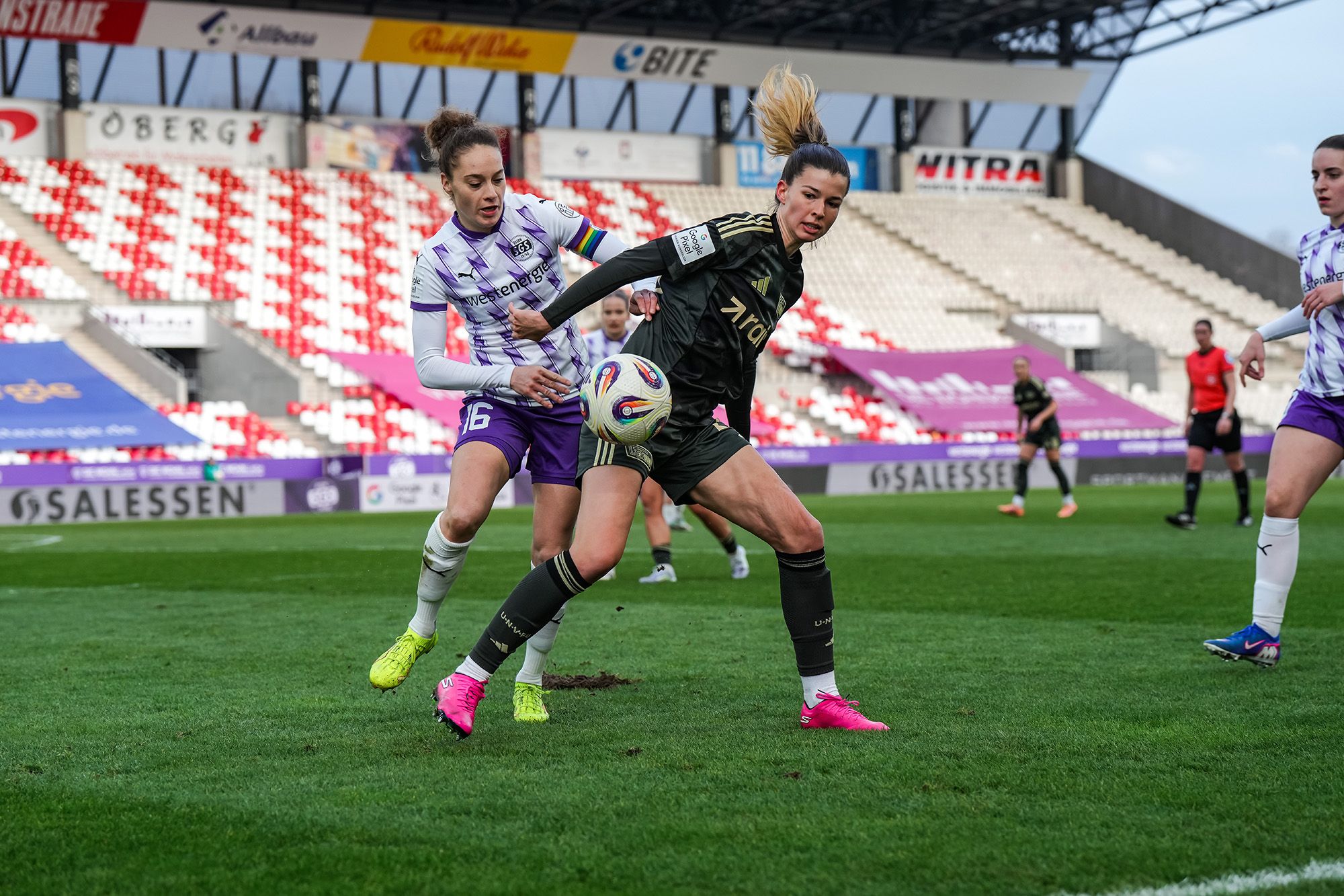 Zwei Frauen im Fußballtrikot kämpfen um den Ball auf einem Sportplatz mit Tribünen im Hintergrund.