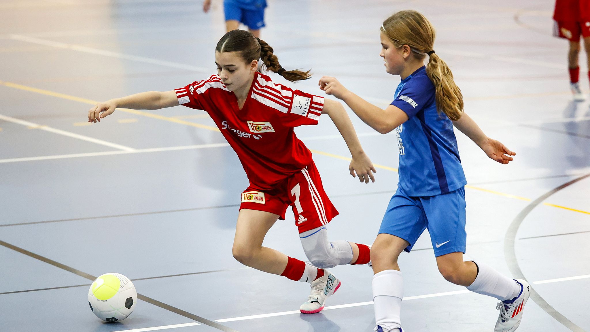 Zwei Mädchen spielen Fußball in der Halle; das Mädchen in Rot dribbelt den Ball, während das in Blau versucht, ihn zu erreichen.