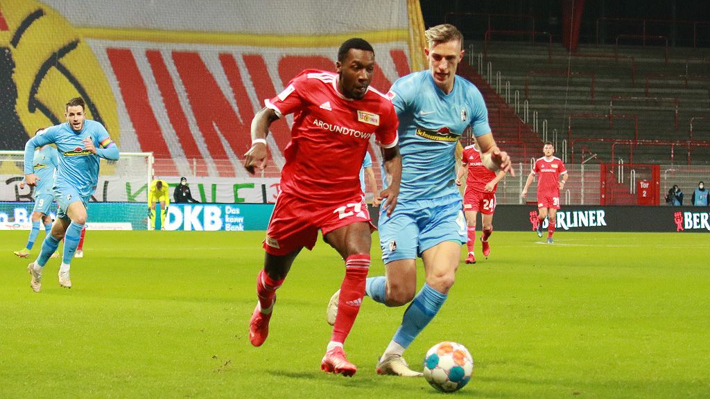 Zwei Fußballspieler im Wettlauf um den Ball auf einem Stadionfeld, im Hintergrund sind Fans und Werbebanner sichtbar.