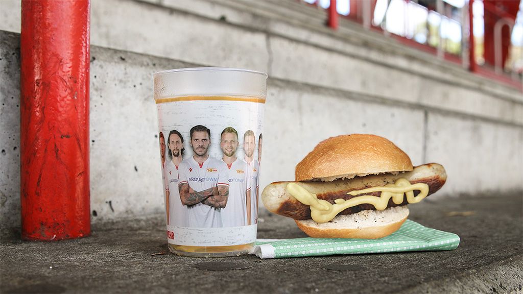 Ein Glas mit einem Mannschaftsbild und ein Hotdog-Sandwich mit Senf liegen auf einer Betonfläche.