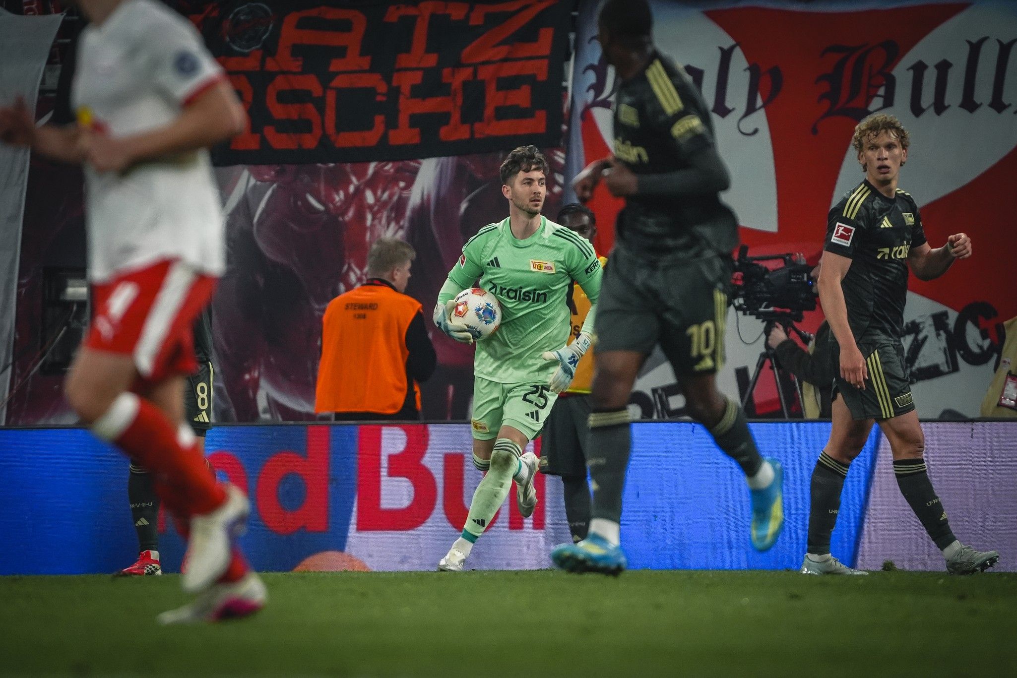 Torwart in grüner Uniform mit Ball, während ein Fußballspiel im Hintergrund stattfindet, umgeben von Fans und Banner.