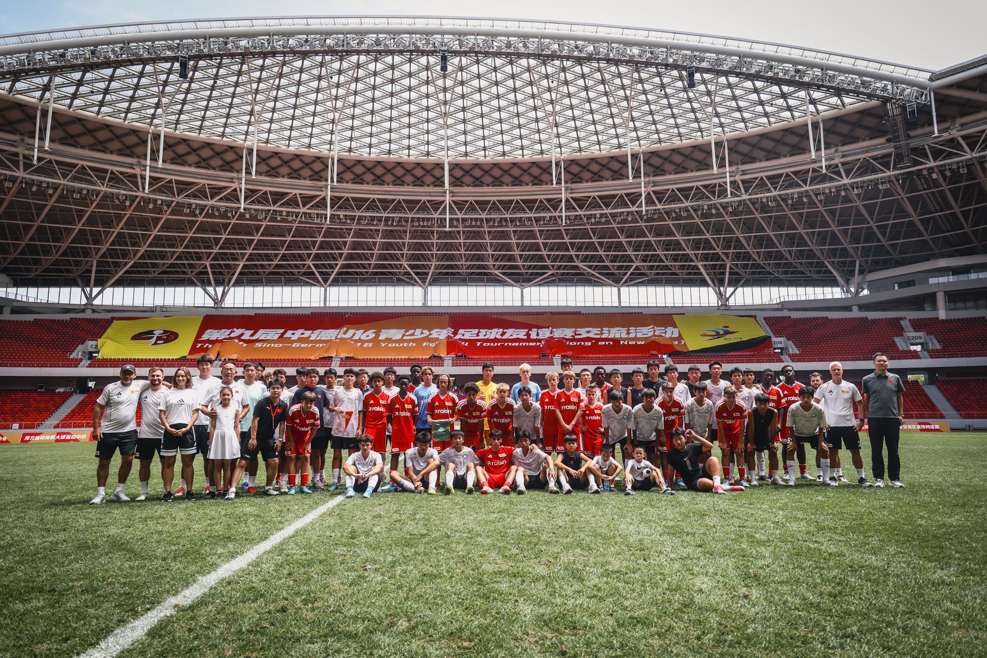 Eine große Gruppe von Menschen in Fußballtrikots steht auf einem Stadionfeld, umgeben von Tribünen und einem Banner im Hintergrund.