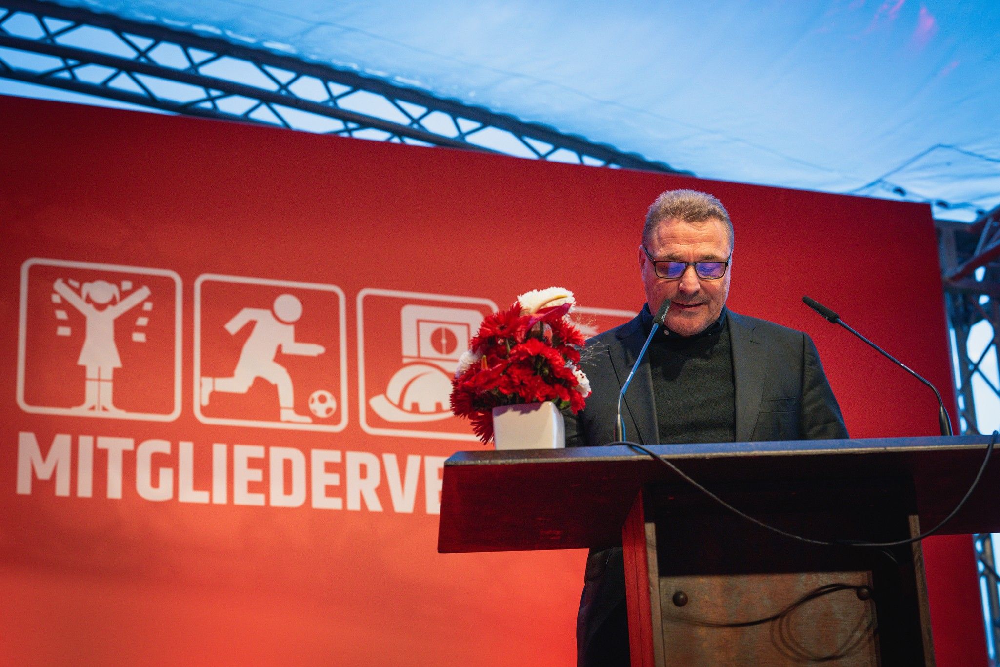 Ein Redner steht an einem Podium, umgeben von Blumen, vor einer rot-weißen Kulisse mit dem Text "MITGLIEDERVERBAND".
