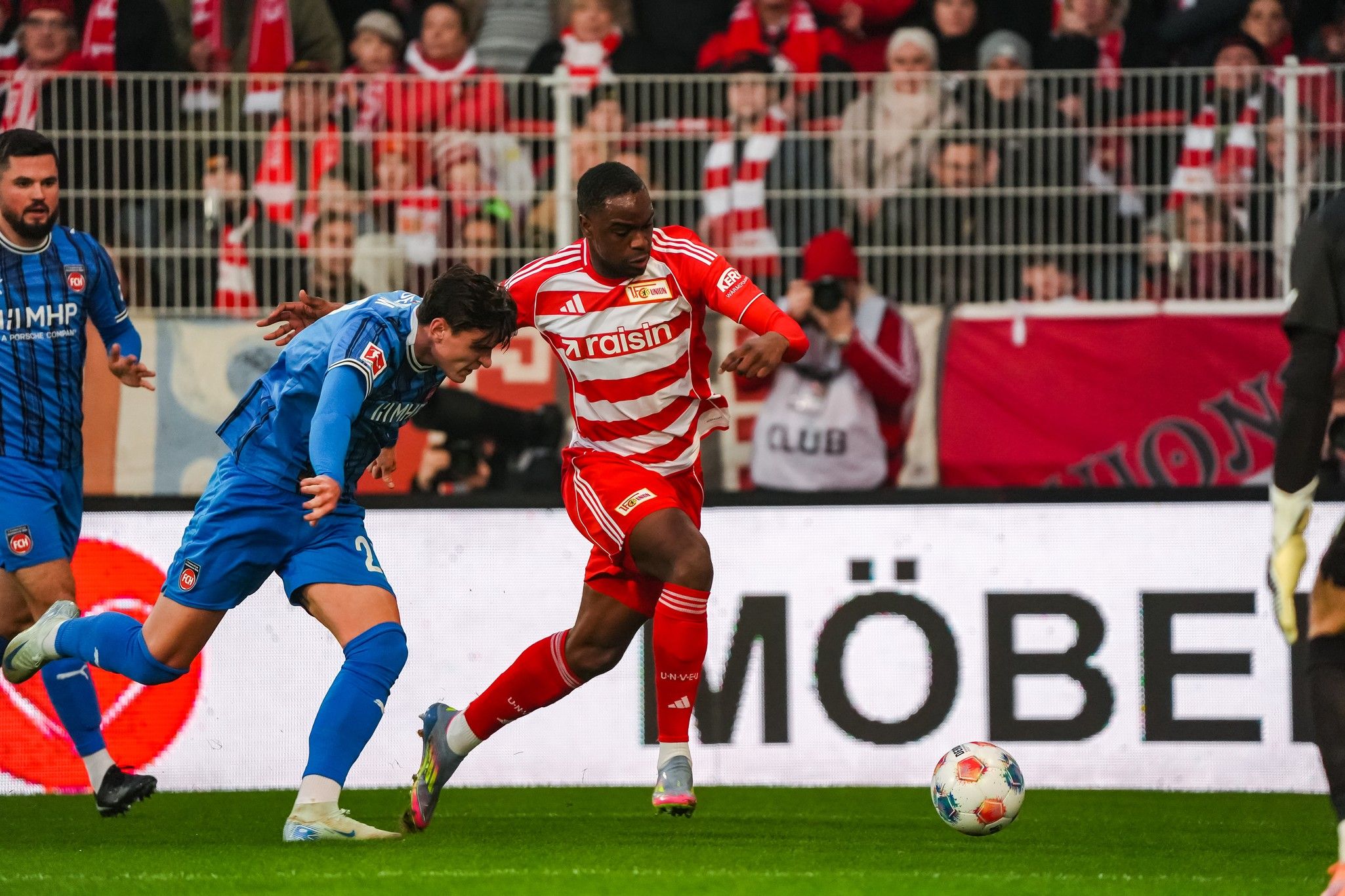 Ein Fußballspieler in rot-weißer Kleidung dribbelt den Ball, während ein blauer Torwart versucht, ihn zu stoppen. Fans im Hintergrund.