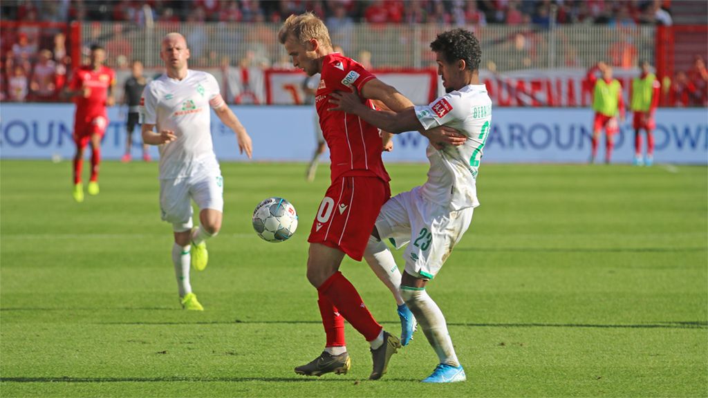 A player in a red jersey approaches the ball while an opponent holds him from behind. In the background, other players and spectators are visible.