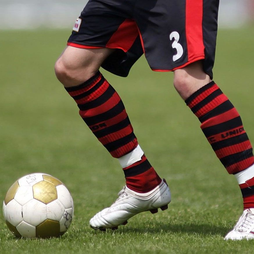 Ein Fußballspieler steht auf dem Rasen und bereitet sich darauf vor, einen Ball zu spielen. Der Spieler trägt ein rot-schwarzes Trikot und Stutzen.