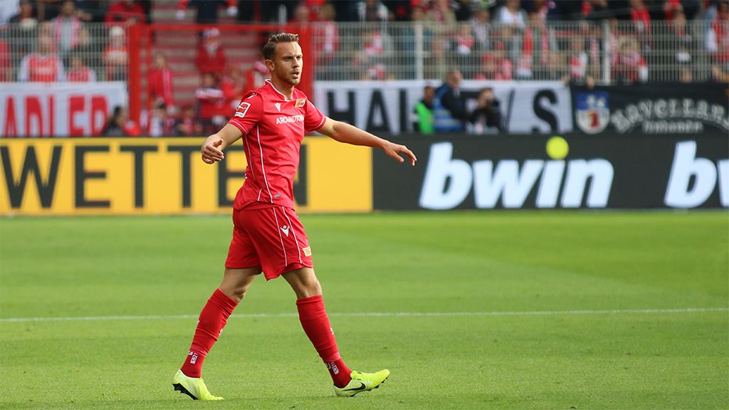 Ein Fußballspieler trägt ein rotes Trikot. Er läuft auf dem Platz. Die Fans im Hintergrund freuen sich und jubeln.