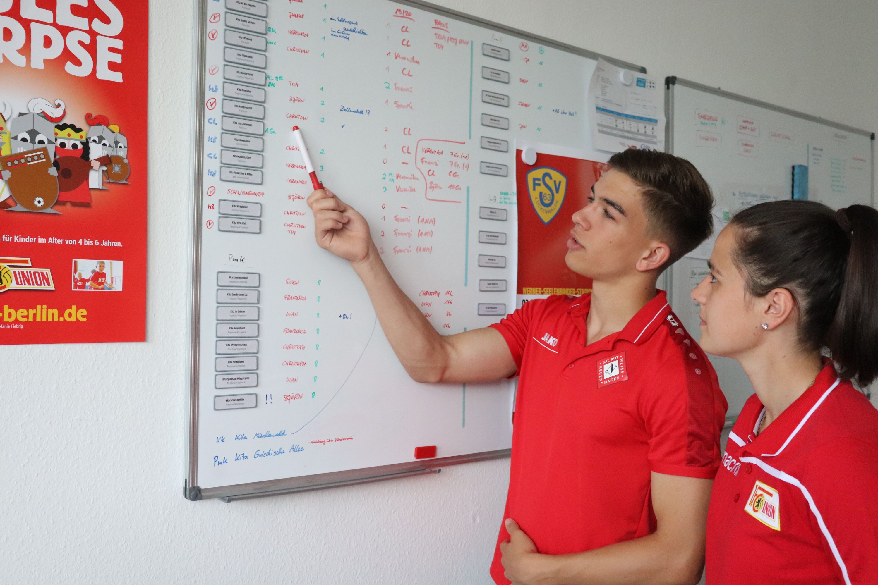 Zwei Personen in roten Sporttrikots stehen vor einem Whiteboard, das mit Informationen und Notizen beschrieben ist.
