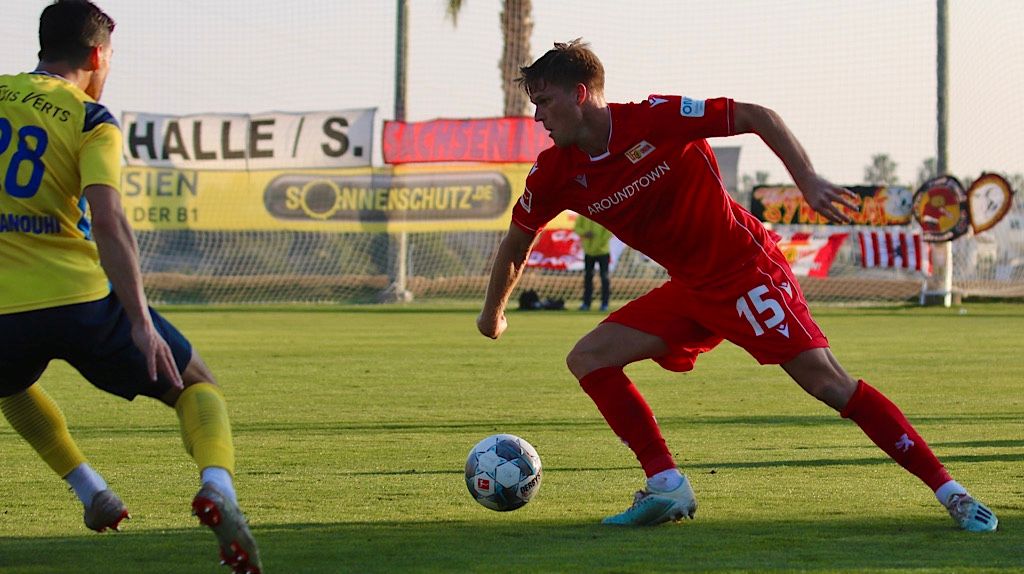 Ein Spieler hat ein rotes Trikot. Er dribbelt den Ball. Ein anderer Spieler hat ein blaues Trikot. Er versucht, den Ball zu stoppen.