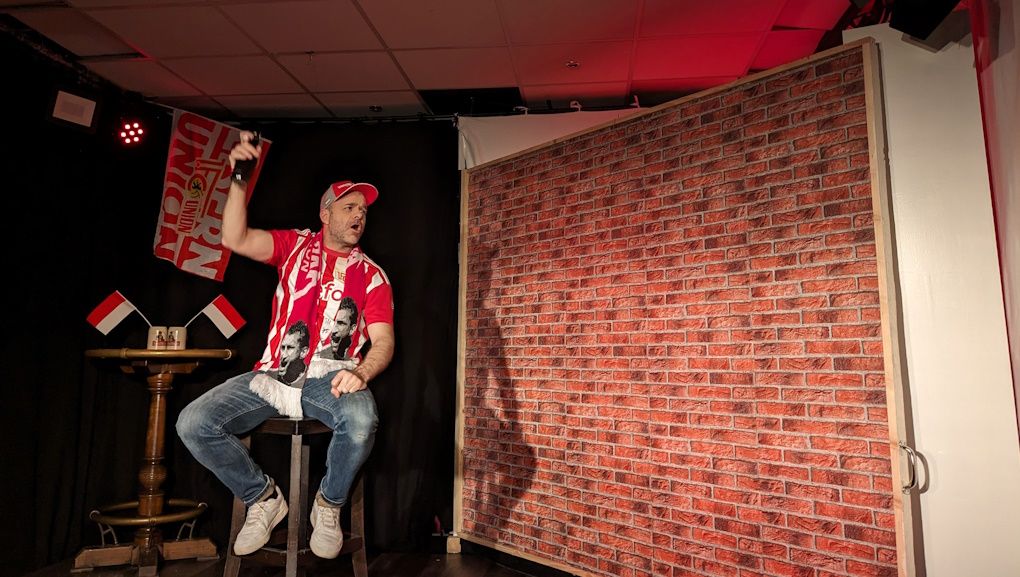 Ein Mann sitzt auf einem Hocker, trägt ein rotes Shirt und einen Hut, und gestikuliert vor einer Backsteinwand.