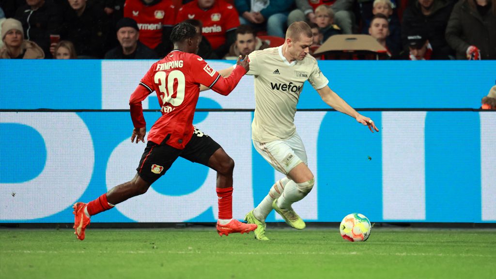 Ein Fußballspieler in weiß dribbelt den Ball, während ein gegnerischer Spieler in rot versucht, ihn zu verteidigen.