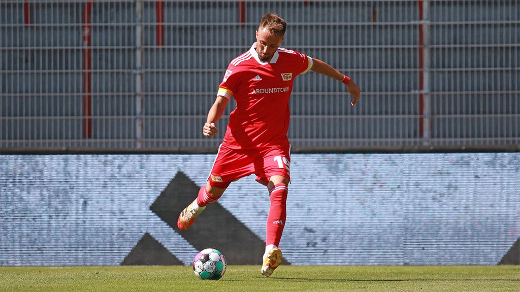 Ein Fußballspieler in rotem Trikot steht auf dem Platz und schlägt einen Schuss auf den Ball.