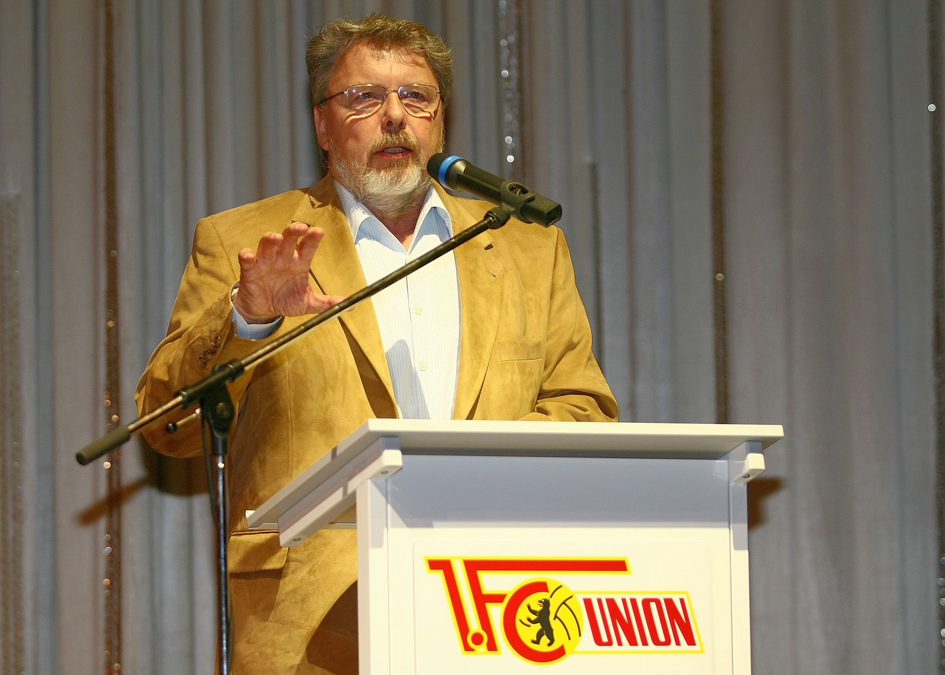 Ein Mann in einem beigen Sakko spricht am Mikrofon vor einer Bühne mit einem Banner von 1. FC Union.
