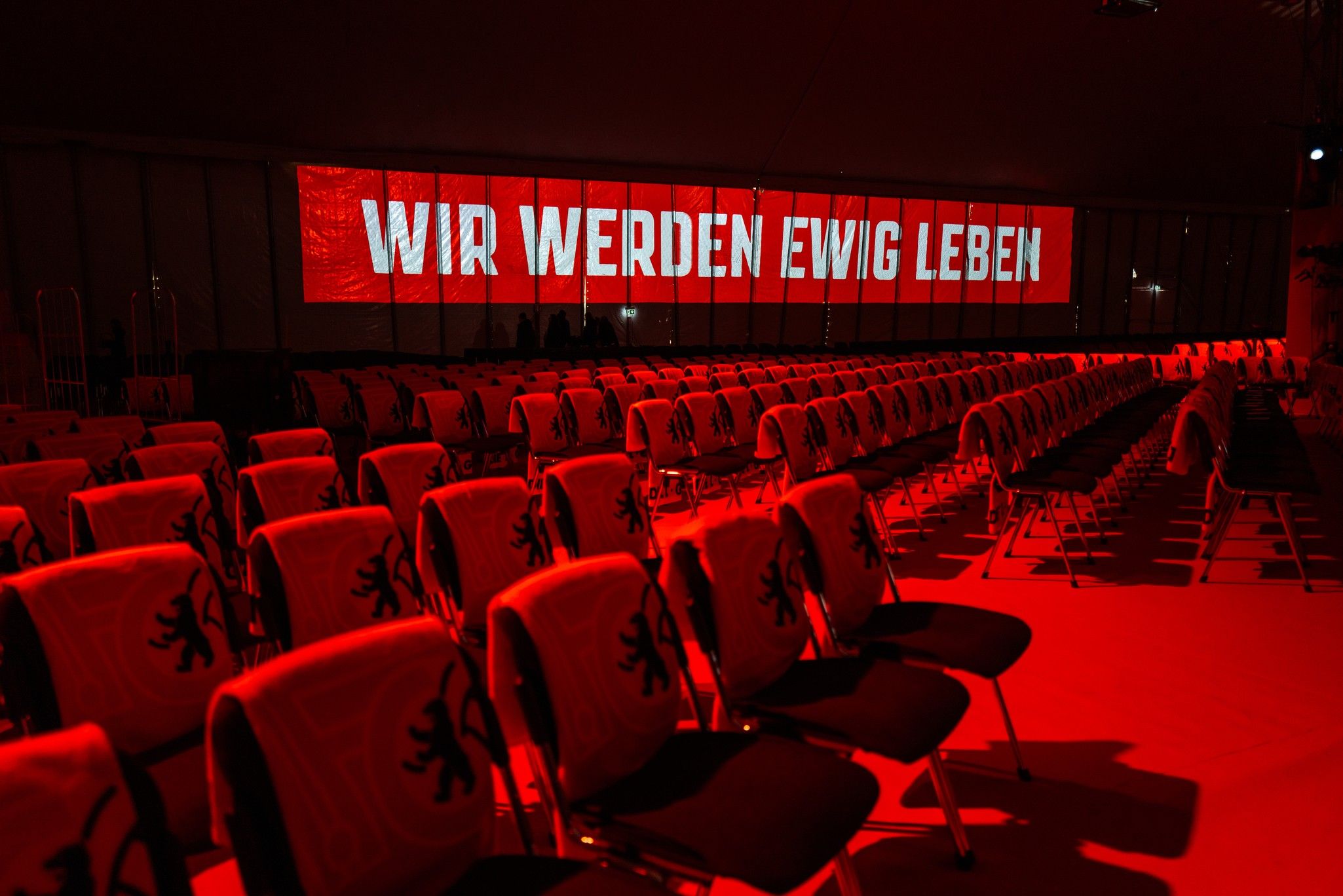 Stuhlreihen in einem rot beleuchteten Raum mit einem Banner, auf dem „WIR WERDEN EWIG LEBEN“ steht.
