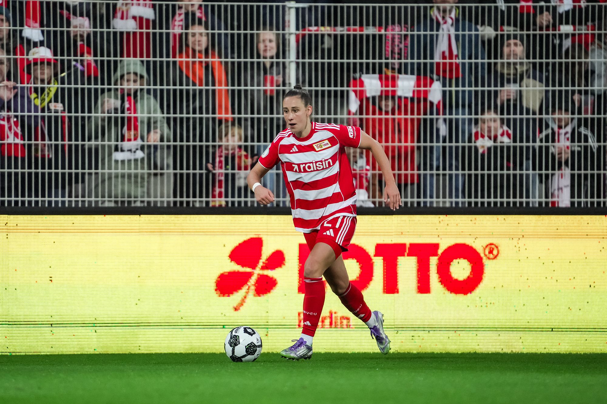 Spieler in rot-weiß gestreiftem Trikot dribbelt den Ball auf einem Fußballfeld, Fans im Hintergrund jubeln.