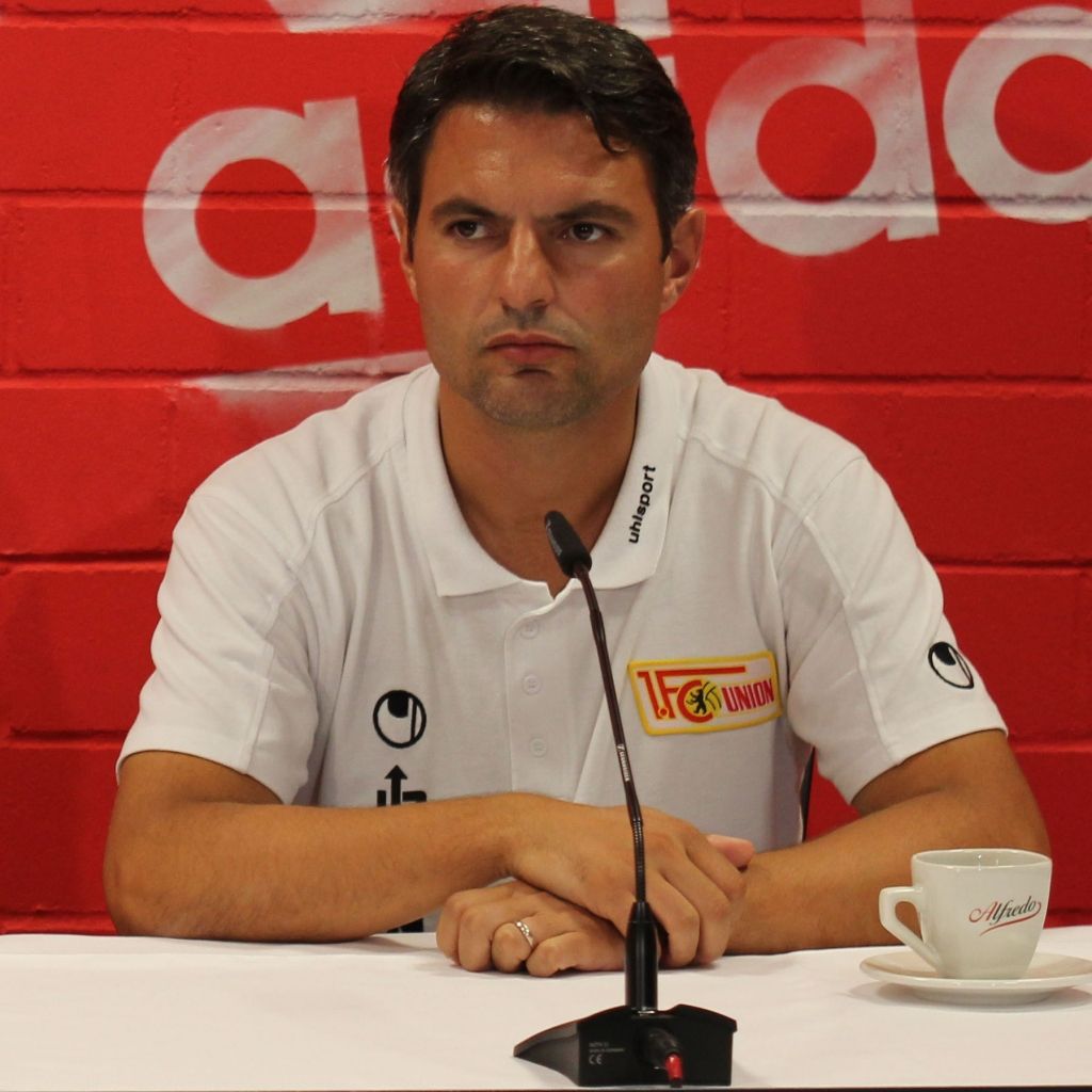 Ein Mann in einem weißen Poloshirt sitzt an einem Tisch mit einem Mikrofon und einer Tasse, vor einer roten Wand mit Logos.