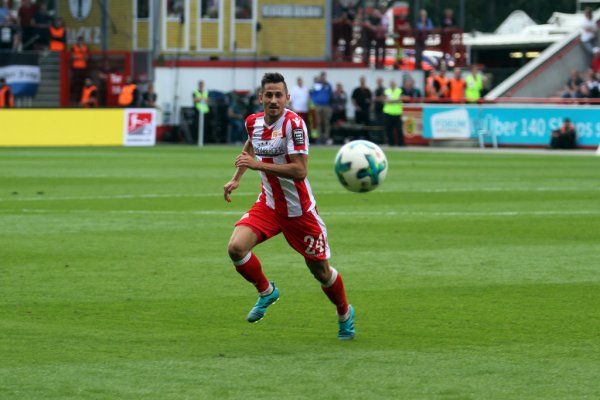 Ein Fußballspieler in roter und weißer Uniform verfolgt den Ball auf einem Spielfeld während eines Spiels.
