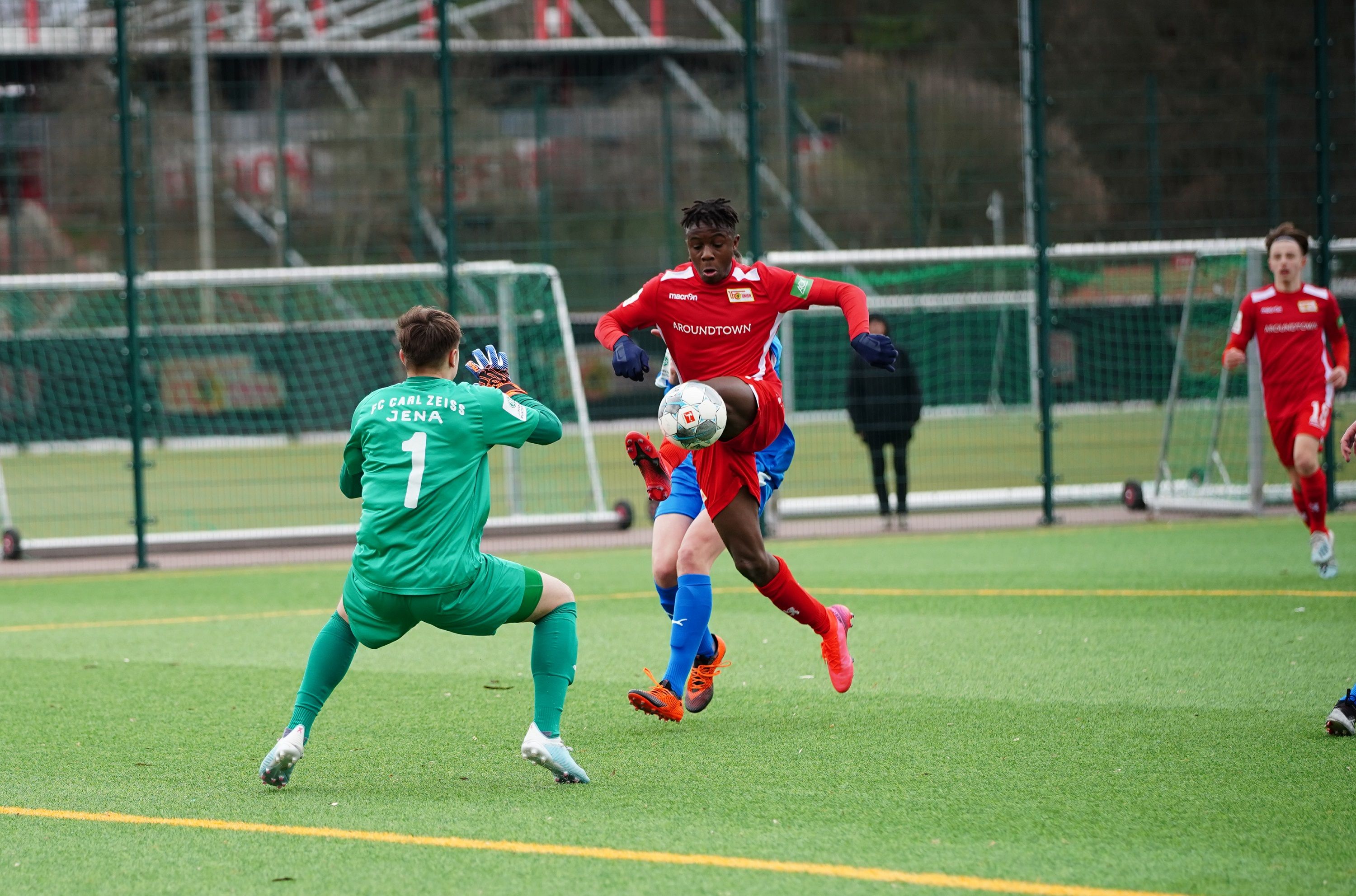 Fußballspiel mit einem Angreifer in rotem Trikot, der auf den Torhüter in grün zustürmt, während er den Ball kontrolliert.