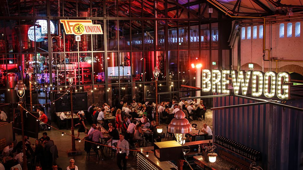 Blick in eine lebhafte Brauerei mit Gäste in einem großen, offenen Raum, festlicher Beleuchtung und modernen Brauanlagen.