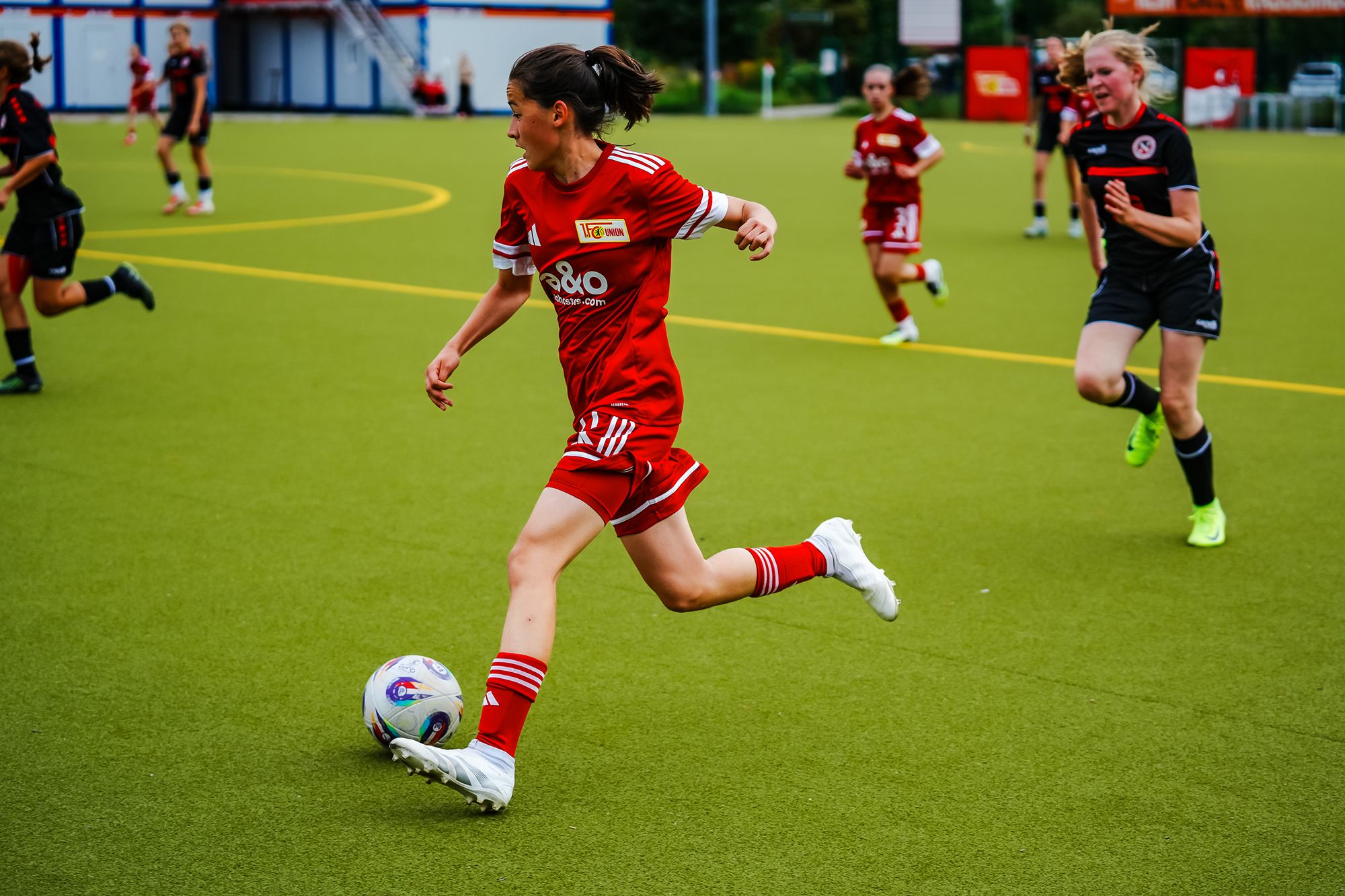 Eine Fußballspielerin in rotem Trikot rennt über das Spielfeld und verfolgt einen Ball, während eine Gegenspielerin im Hintergrund ist.