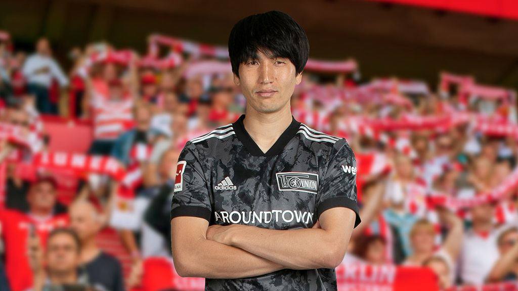 Ein Fußballspieler in einem schwarzen Trikot steht mit verschränkten Armen vor einer jubelnden Menge im Stadion.
