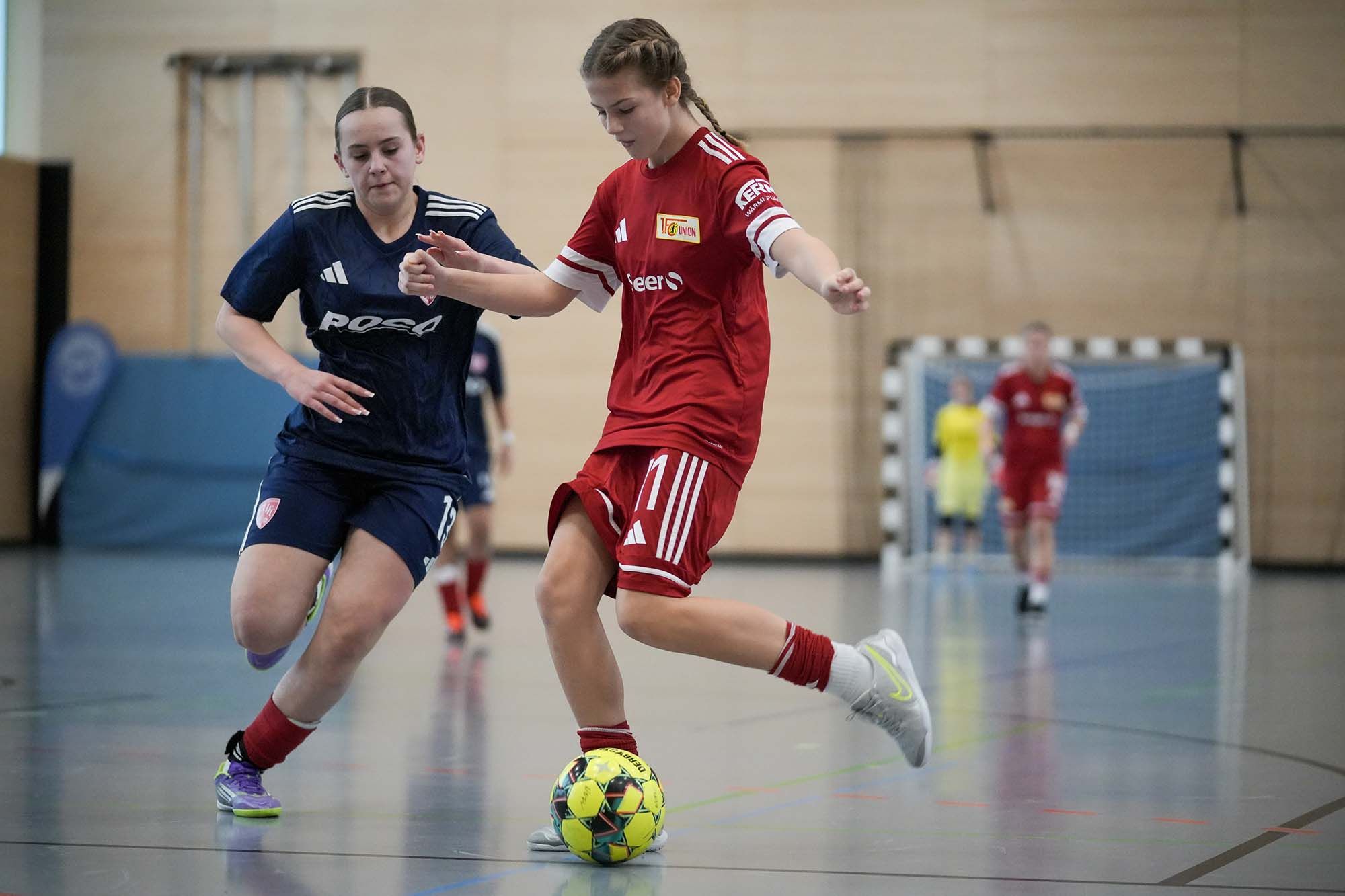 Zwei Mädchen im Fußballspiel in einer Halle, das eine in rotem Shirt dribbelt, das andere in blauem Shirt versucht, den Ball zu erobern.