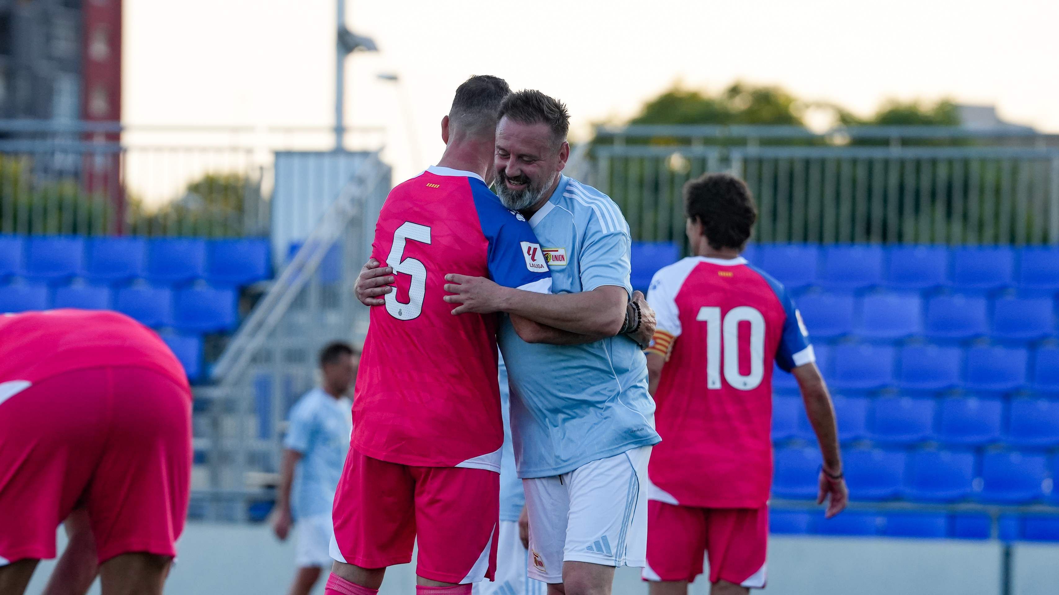 Zwei Fußballspieler umarmen sich nach dem Spiel. Der eine trägt ein pinkes Trikot mit der Nummer 5. Der andere trägt ein blaues Trikot.