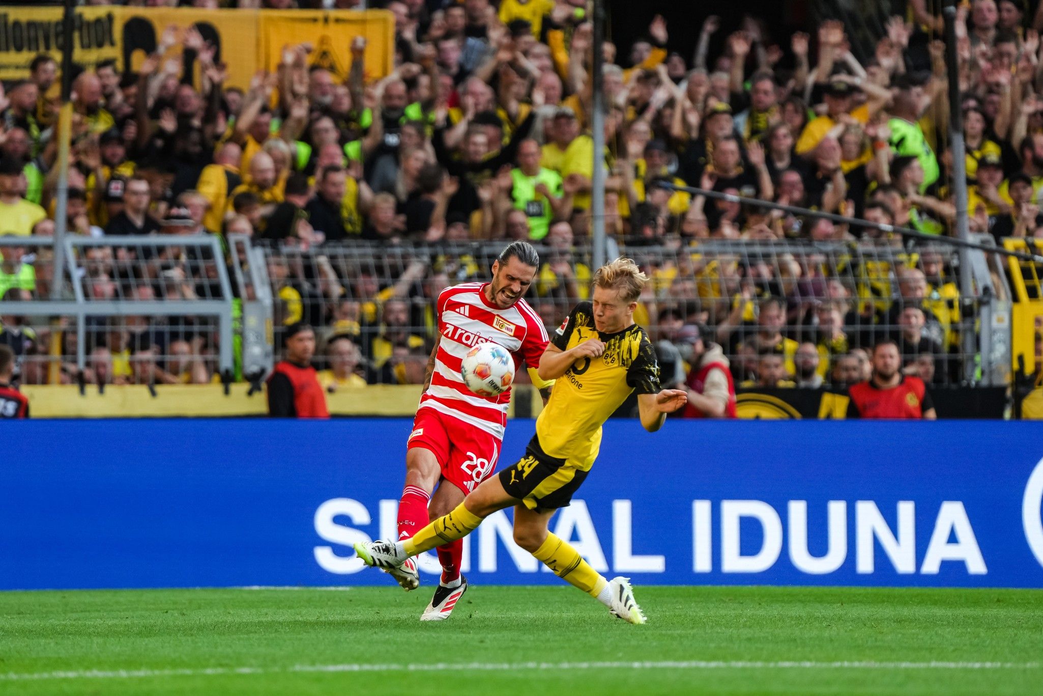 Ein Fußballspieler in gelber Uniform tritt den Ball, während ein gegnerischer Spieler in rot-weißer Uniform ihn bedrängt. Hintergrund mit jubelnden Fans.
