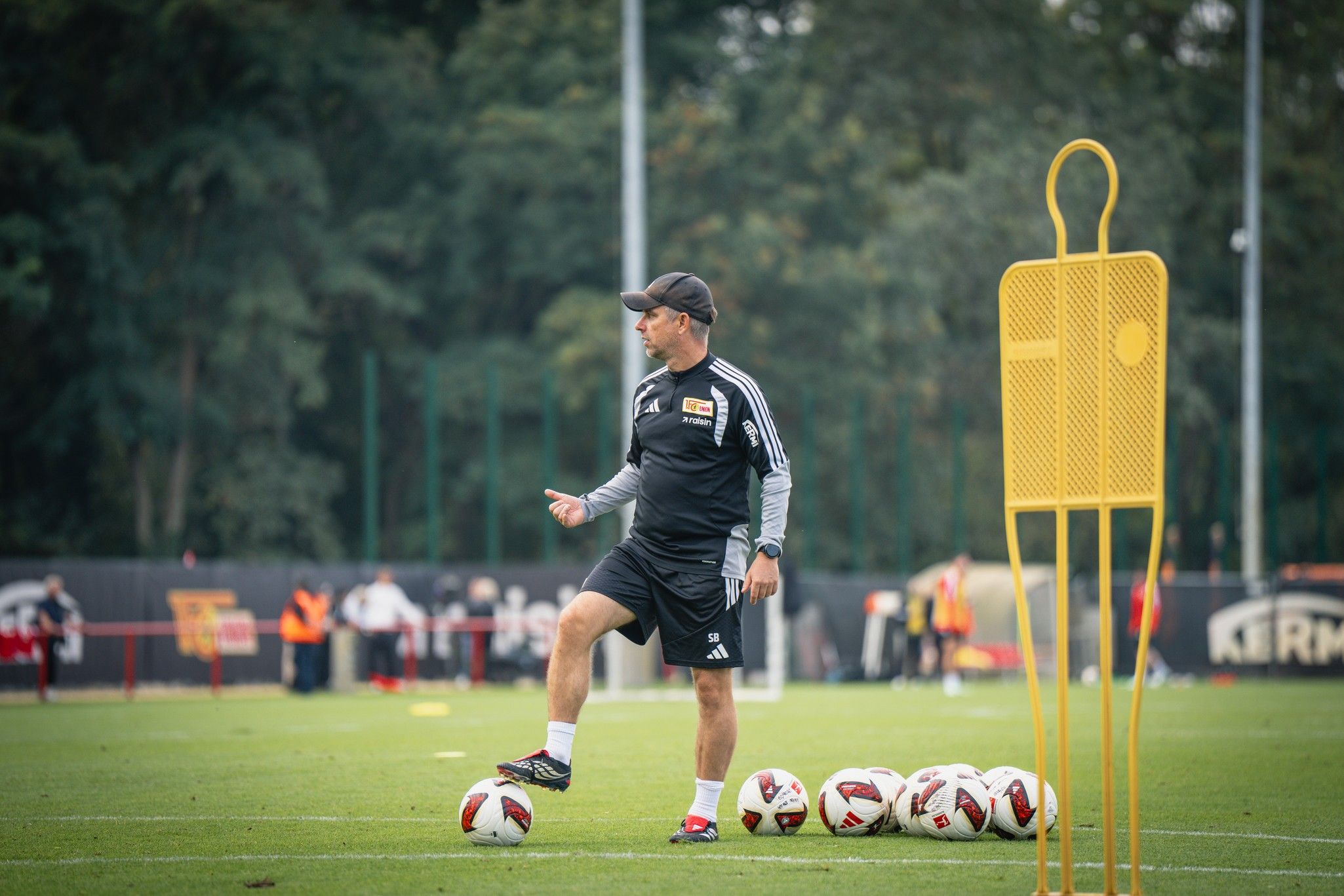Ein Trainer leitet das Fußballtraining auf einem Rasenplatz, umgeben von Trainingsutensilien und Spielern im Hintergrund.