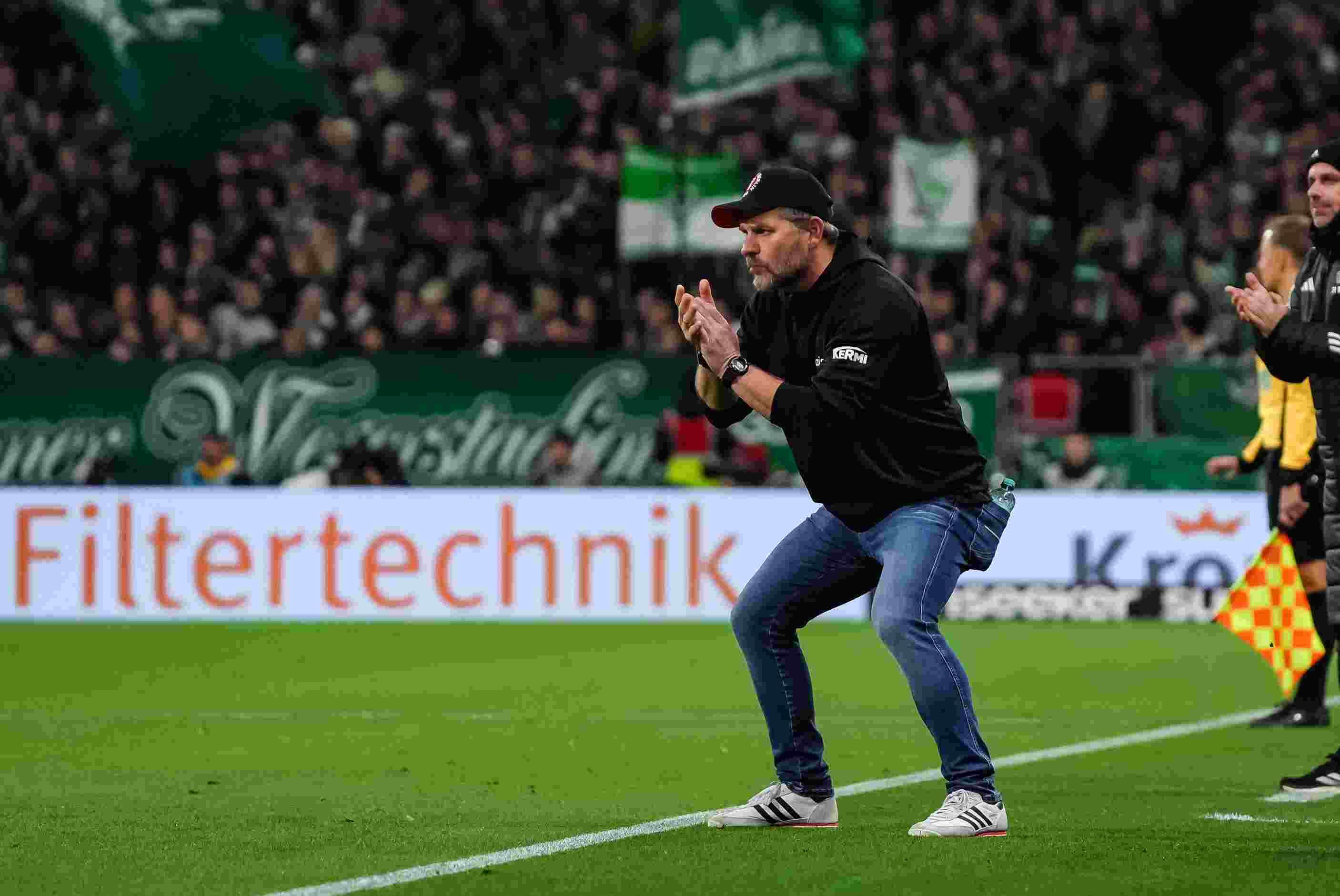 Ein Trainer applaudiert und gibt Anweisungen während eines Fußballspiels auf dem Platz. Im Hintergrund sind Fans und Banner zu sehen.