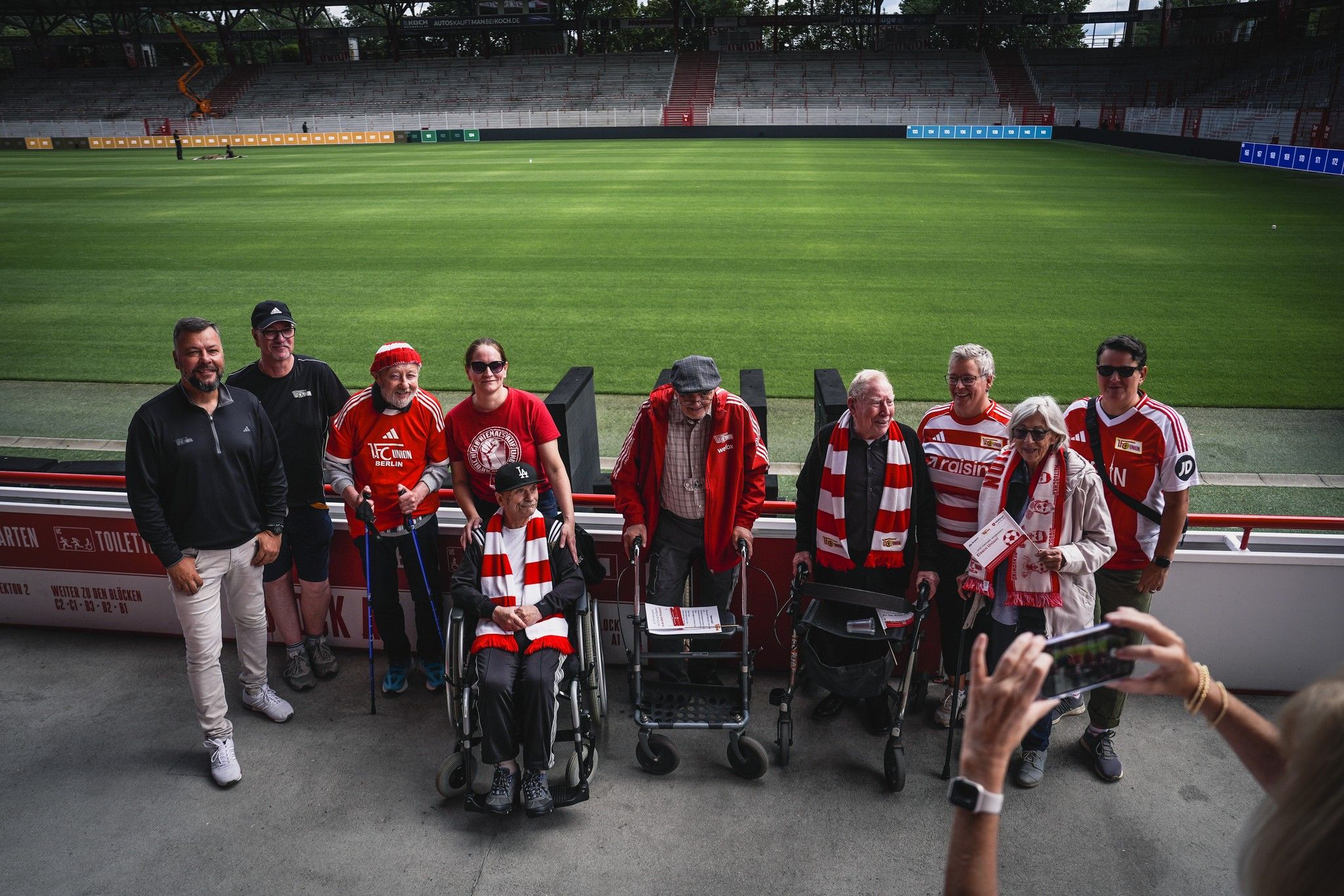 Eine Gruppe von älteren Menschen in roten T-Shirts und Schals posiert auf einem Fußballfeld, einige mit Rollatoren.