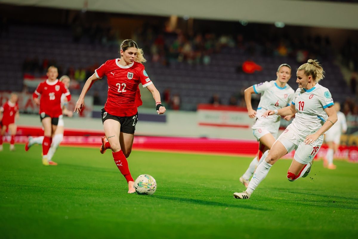 Eine Spielerin in rotem Trikot dribbelt den Ball, während eine Gegnerin in weißem Trikot sie verfolgt. Stadionatmosphäre im Hintergrund.