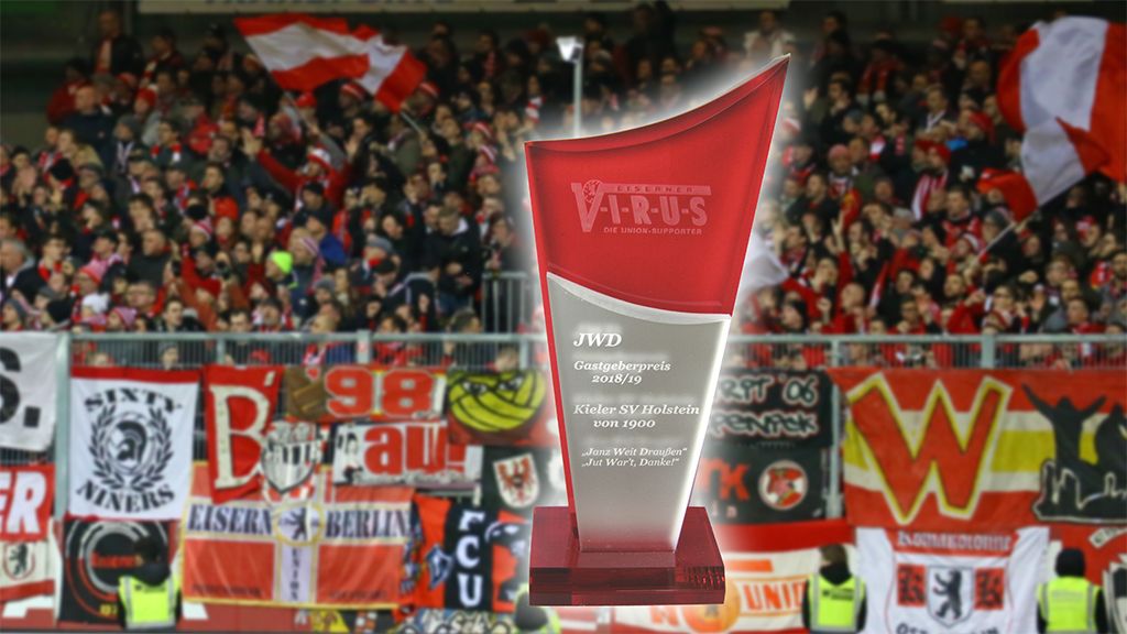Pokal mit der Aufschrift "Gastgeberpreis 2018/19" vor einer jubelnden Menschenmenge mit Fankultur in einem Stadion.