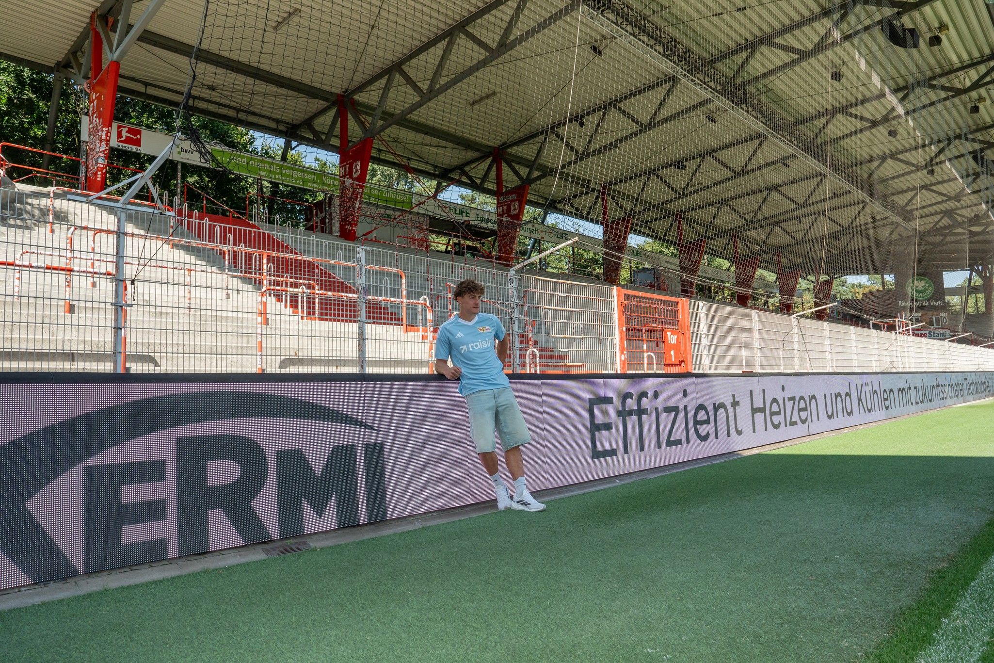 Junger Mann in hellblauer Sportkleidung lehnt an einer Werbebande in einem Stadion mit leeren Sitzplätzen.