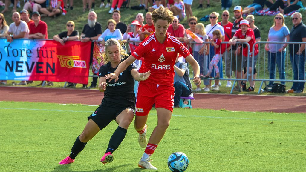 Eine Fußballspielerin in rotem Trikot dribbelt den Ball, während eine Gegnerin im schwarzen Trikot versucht, sie zu stoppen. Zuschauer im Hintergrund.