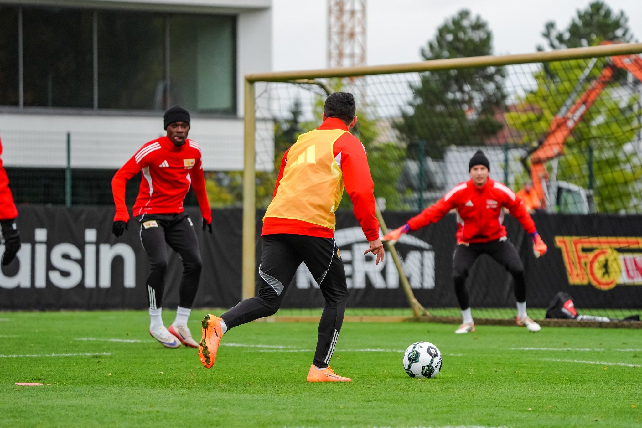 Spieler in roten Trainingsanzügen üben Fußball auf einem Rasenplatz, während ein Spieler den Ball dribbelt.