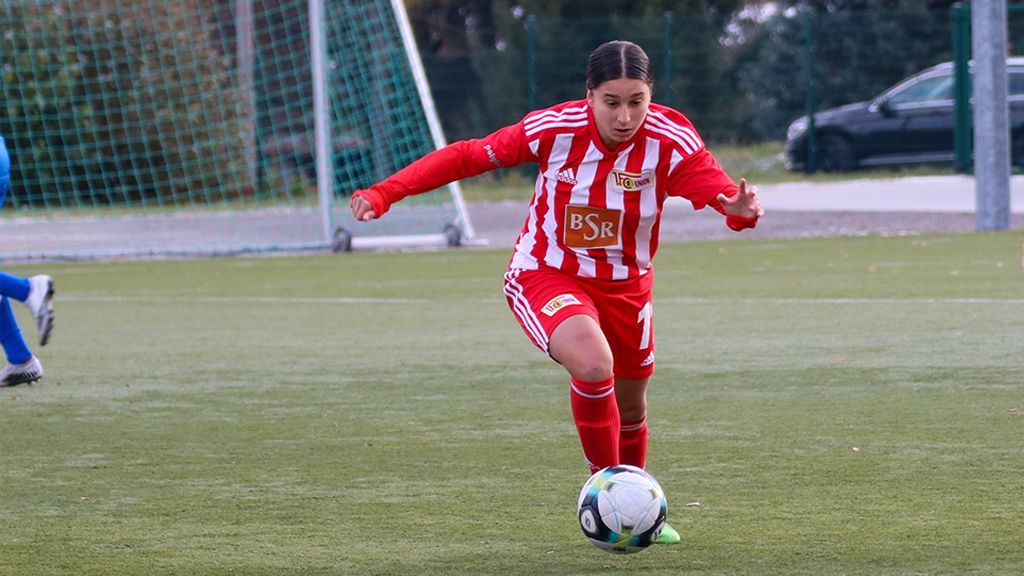 Eine Fußballspielerin in rot-weißer Trikott sieht konzentriert aus, während sie den Ball auf einem Kunstrasenfeld dribbelt.