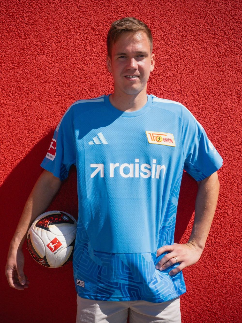 Ein junger Mann in einem blauen Fußballtrikot steht an einer roten Wand und hält einen Fußball in der Hand.