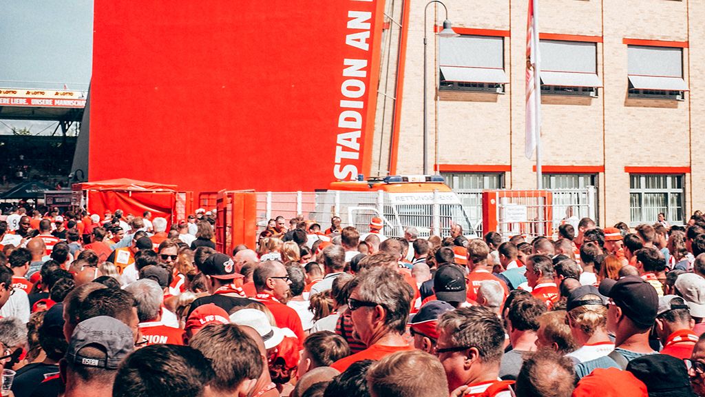Menschenmenge vor einem Stadion, gekleidet in rote Kleidung und mit Fahnen, umgeben von Gebäuden und Stadioninfrastruktur.