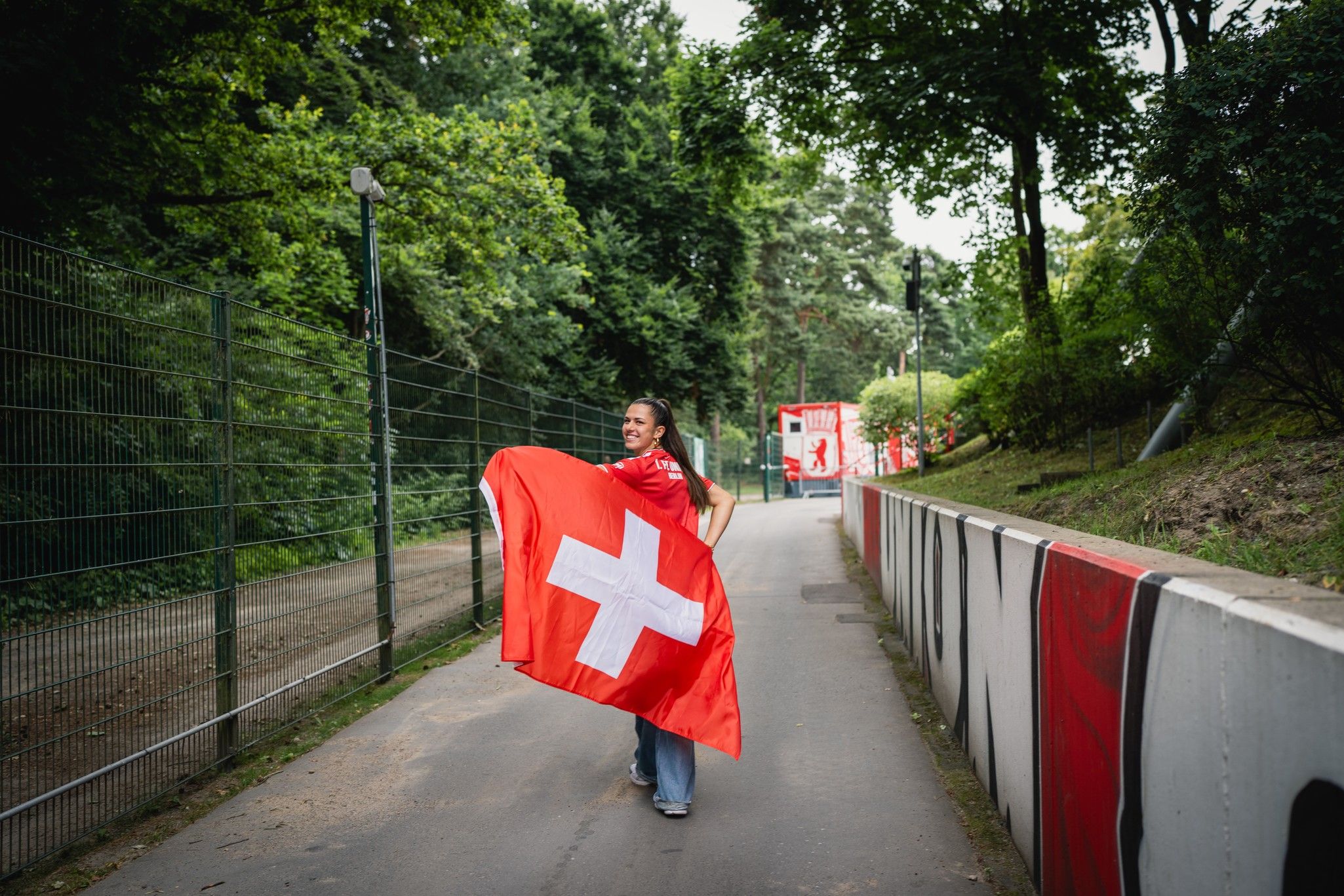 Eine Person trägt eine große Schweizer Flagge und geht auf einem Weg entlang Bäume und Zäune.