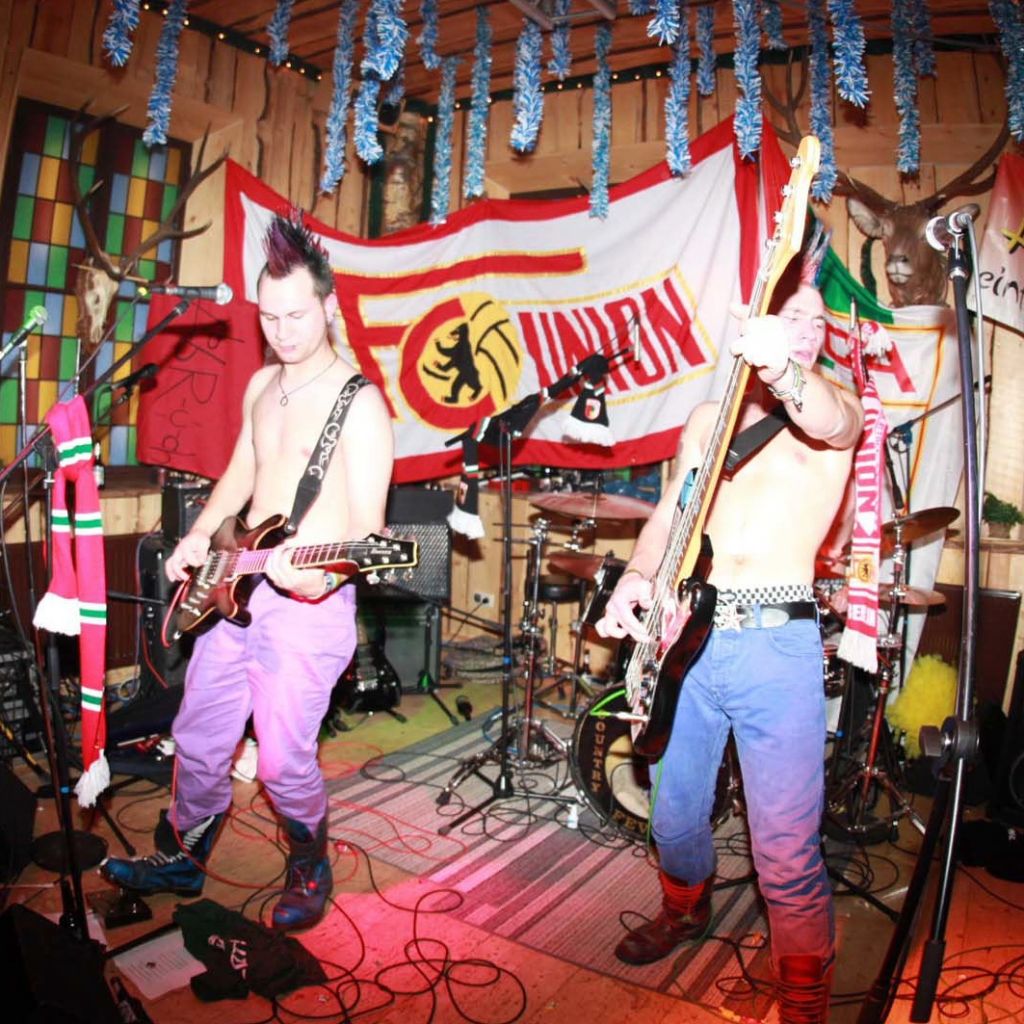 Zwei Musiker mit bunten Haaren und kontrastreichen Outfits spielen live auf einer Bühne, umgeben von Instrumenten und Dekoration.