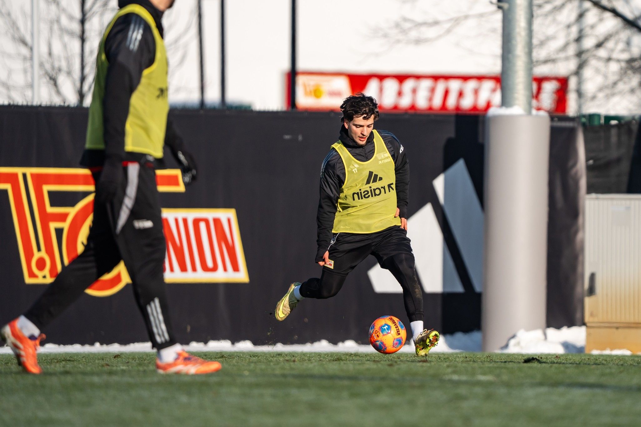 Ein Fußballspieler in gelbem Trainingsleibchen schießt einen Ball auf einem Rasenplatz, im Hintergrund sieht man eine andere Person.