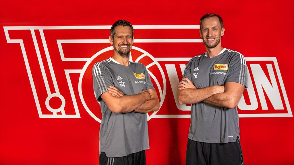 Zwei Männer in grauen Sport-T-Shirts stehen lächelnd vor einem roten Hintergrund mit dem Logo eines Vereins.