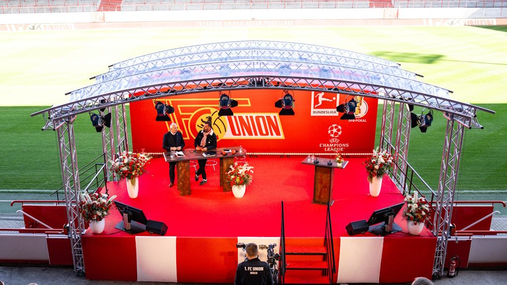 Bühne mit Tisch und Stühlen, dekoriert mit Blumen, auf einem Sportplatz im Stadion, im Hintergrund Union Berlin-Logo.