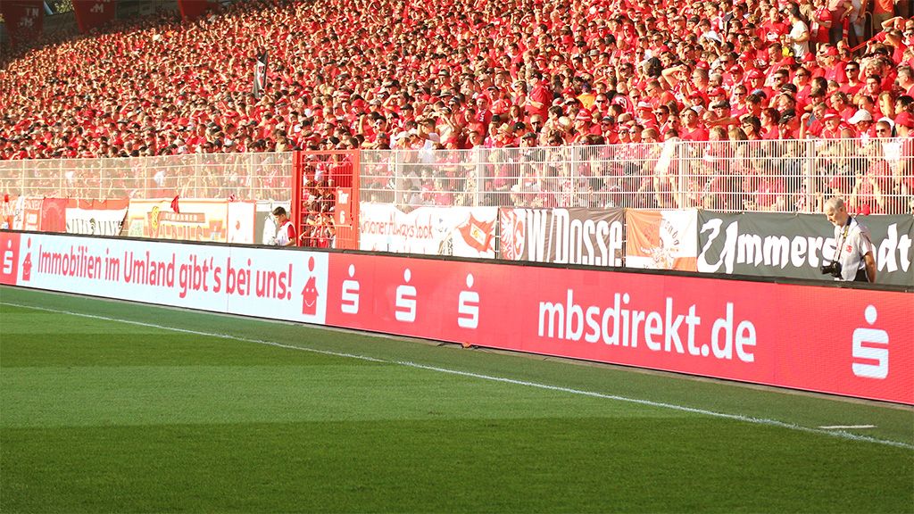Ein großer Teil der Stadiontribüne mit jubelnden Fans in roten T-Shirts und verschiedenen Bannern.
