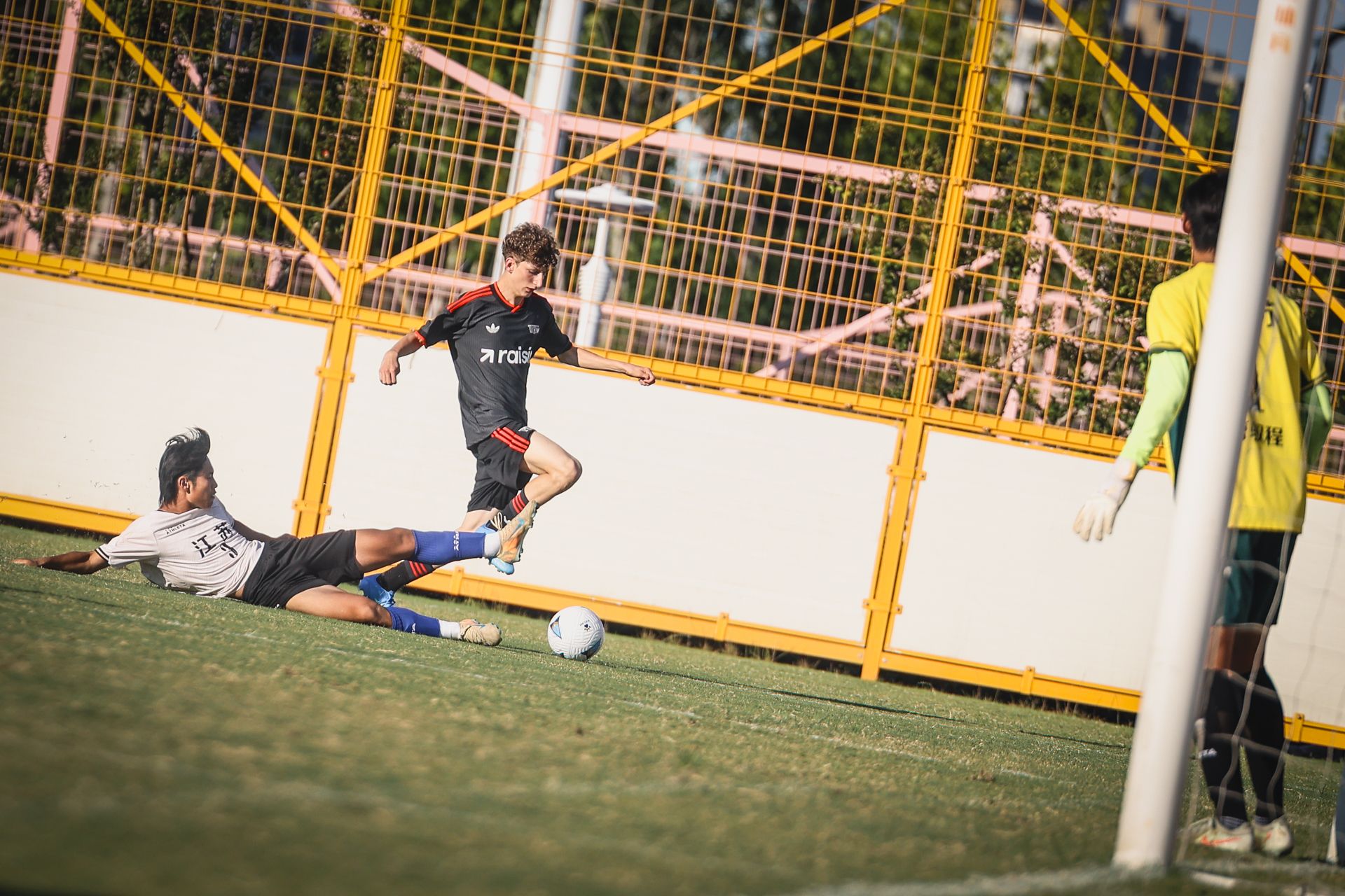 Ein Fußballspieler springt über einen am Boden liegenden Gegner, während ein Torwart am Tor steht.