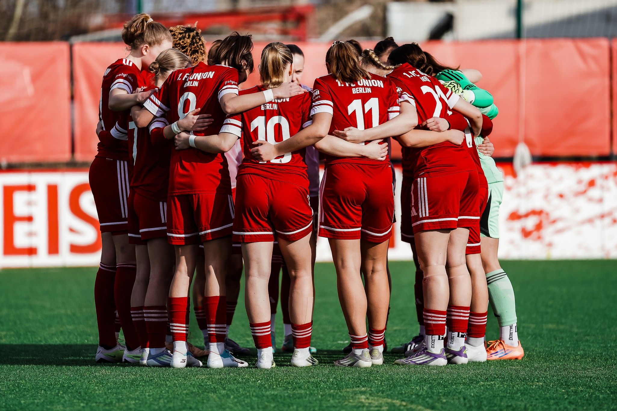 Spielerinnen eines Frauenteams in roten Trikots umarmen sich im Kreis auf dem Spielfeld vor einem Spiel.