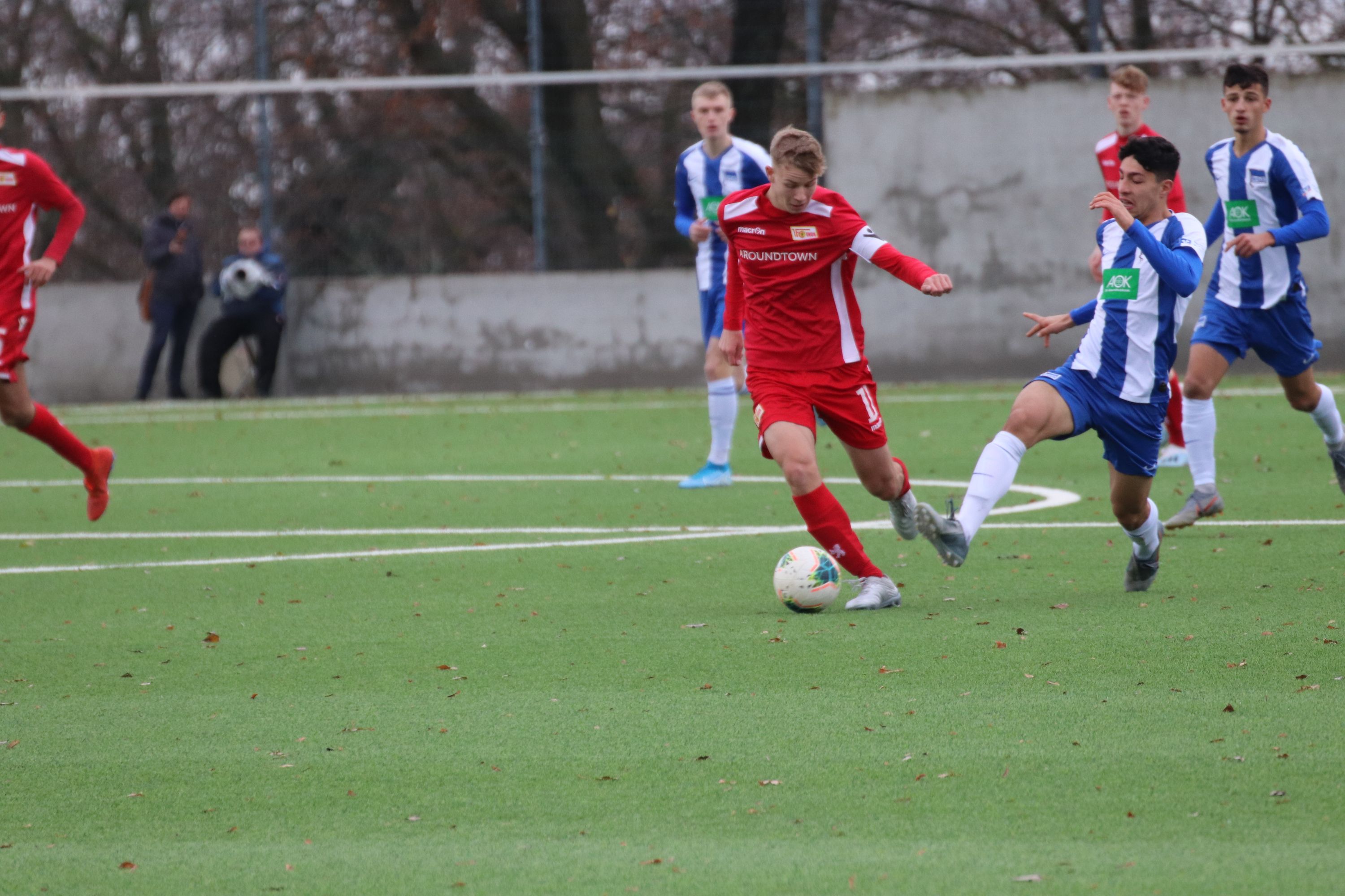 Spielszene eines Fußballspiels, in dem ein Spieler in roter Kleidung einen Gegner in blauer Kleidung am Ball hindert.