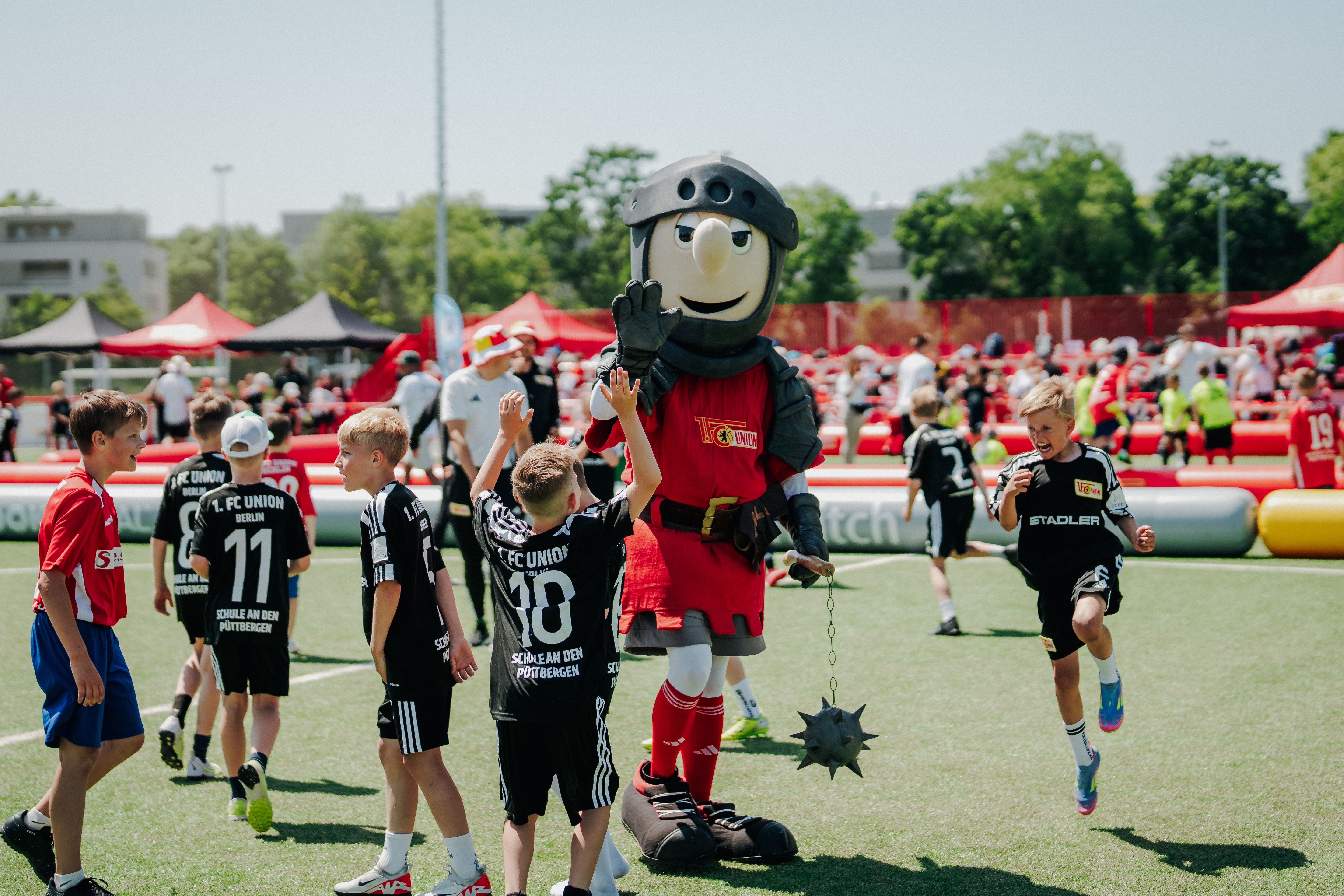 Kinder in Fußballtrikots spielen auf einem Sportplatz, während ein Maskottchen in Ritterkostüm lächelt und mit ihnen interagiert.